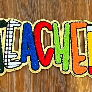 May include: A colorful felt banner with the word "TEACHER" in gold outlined letters. Each letter is a different color: green, blue, red, green, orange, and yellow. The "E" has a small red pencil drawn on it.