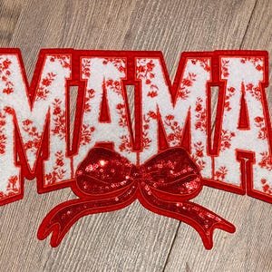 May include: Red felt applique with the word "MAMA" in white letters with a red sequined bow. The letters have a white floral pattern.