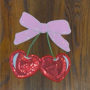 May include: A pair of red cherries with a pink bow on top. The cherries and bow are made of sequins.
