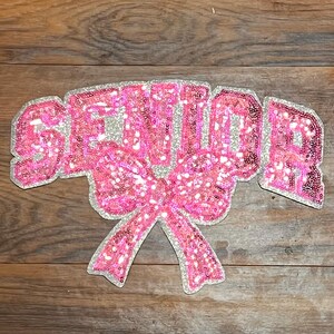 May include: Pink and silver sequined iron-on patch with the word "Senior" and a large pink bow.