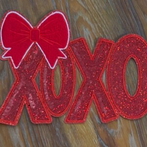 May include: Red glitter XOXO with a red bow on top. The letters are outlined in white.