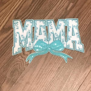 May include: A blue and white fabric patch with the word "MAMA" in large, white letters with a blue bow in the center. The patch is decorated with a small floral pattern.