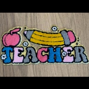 May include: A colorful, glittery patch with the word "TEACHER" in bubble letters. The design includes a pink apple, a yellow pencil, and small flower accents. The patch is likely for a teacher.
