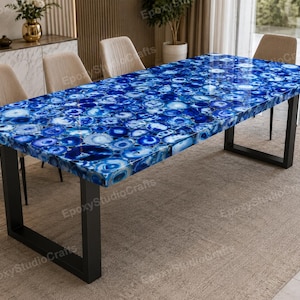 Puede incluir: Una mesa de comedor rectangular con un tablero pulido compuesto por numerosas rodajas de ágata azul con patrones concéntricos. La mesa está sostenida por dos patas de metal negro en forma de U, que proporcionan una base estable a la piedra.