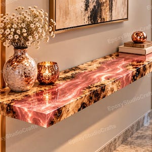 Puede incluir: Una consola flotante montada en la pared con un tablero de madera de borde natural y una incrustación central de resina rosa brillante. La mesa sostiene un jarrón moteado con flores blancas, un portavelas metálico y libros apilados.