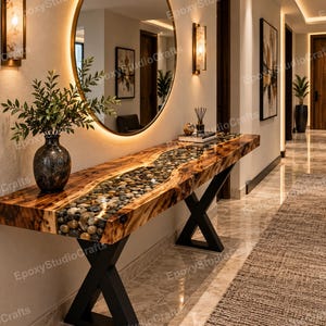 Op de afbeelding: Een consoletafel met een natuurlijke houten rand en een centrale inleg gevuld met gladde, veelkleurige rivierstenen ingebed in heldere epoxyhars. De tafel wordt ondersteund door zwarte, X-vormige metalen poten en staat tegen een muur.