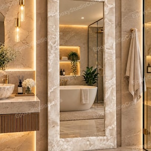 May include: A tall, rectangular floor mirror featuring a wide frame with a beige and white marbled pattern. The mirror surface reflects a bathroom interior, including a white freestanding bathtub, a green plant, and tiled walls.