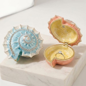 May include: Two seashell-shaped trinket boxes. One is closed, featuring a light blue exterior with a spiral of clear rhinestones. The other is open, revealing a pale yellow interior and a peach-colored exterior, containing a silver ring with a stone.