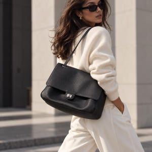 May include: A black leather shoulder bag with a textured finish, a rectangular flap, and a silver-toned metal turn-lock closure. The bag features a single thin shoulder strap and is worn over the shoulder, showcasing a structured design.
