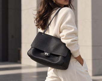 May include: A black leather shoulder bag with a textured finish, a rectangular flap, and a silver-toned metal turn-lock closure. The bag features a single thin shoulder strap and is worn over the shoulder, showcasing a structured design.
