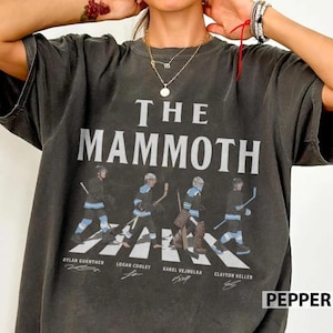 May include: A charcoal gray short-sleeve t-shirt featuring the text THE MAMMOTH in large white letters above four hockey players on a crosswalk. Below each player are the names Dylan Guenther, Logan Cooley, Karel Vejmelka, and Clayton Keller.
