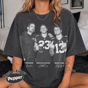 May include: A charcoal grey short-sleeve t-shirt featuring a black and white graphic of Kyle Shanahan, Christian McCaffrey, and Brock Purdy. The design includes their names, jersey numbers, and signatures. The shirt has a vintage, washed-out look.