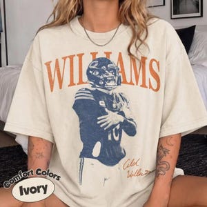 May include: An ivory short-sleeve t-shirt featuring the orange text WILLIAMS in a bold arched font. Below the text is a blue graphic of a football player in a helmet and jersey with the number 10, accompanied by a signature in orange script.