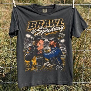 May include: A charcoal gray short-sleeve t-shirt featuring a graphic print titled BRAWL on Speedway. The design shows two race car drivers in racing suits, stadium lights, and text reading Tempers Run Higher at 200 MPH, NASCAR, and Racers Rivals Legends.