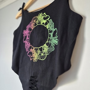 May include: Black tank top with a colorful mushroom design. The graphic features a circular arrangement of mushrooms in shades of pink, yellow, and green. The tank top has a distressed hem and is hanging on a wooden hanger.