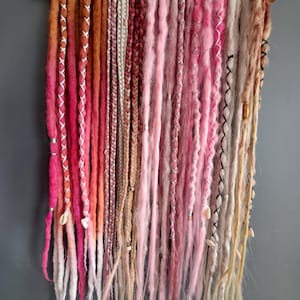 May include: A collection of colorful dreadlocks in shades of pink, brown, and white, hanging from a wooden hanger. Some dreadlocks are adorned with silver accents and small shells. The color palette includes a gradient effect.