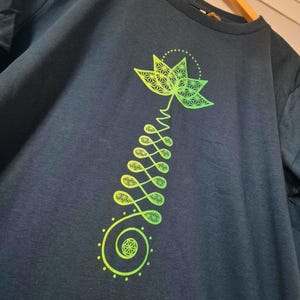 Puede incluir: Sudadera de color verde azulado oscuro con un diseño de flor de loto en verde neón y amarillo. El gráfico incluye una flor de loto detallada, una espiral y formas de hojas conectadas. La sudadera se muestra en una percha de madera.