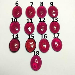 May include: A collection of thirteen oval-shaped, faceted, translucent dark pink gemstones arranged in rows. Each stone is labeled with a black number ranging from 6 to 18, indicating individual pieces for identification and selection purposes.