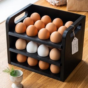 Puede incluir: Un estante de madera negra de tres niveles para almacenar huevos con asas recortadas en los laterales, que contiene veintiún huevos marrones y blancos. Una pequeña etiqueta de papel blanco con texto escrito a mano está atada al lateral.