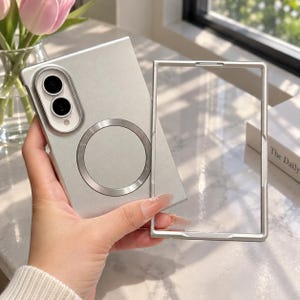 May include: A matte silver smartphone case featuring a circular magnetic ring on the back and a raised camera cutout with two lenses. The set includes a separate, thin, rectangular silver frame that matches the case design.
