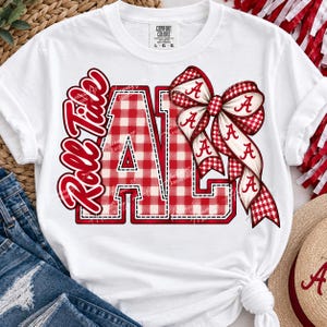 May include: A white short-sleeved t-shirt featuring a red and white gingham pattern graphic that spells ALA with the text Roll Tide in red script. A large red and white gingham bow with the letter A printed on the ribbons is attached to the graphic.
