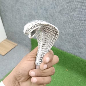 Può includere: Un pomello per bastone in metallo color argento a forma di serpente cobra con il cappuccio spiegato e la bocca aperta. La superficie presenta squame e texture dettagliate su tutta la testa e il collo, con una base cilindrica liscia.