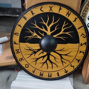 May include: A circular wooden shield featuring a black and natural wood finish. The centre has a black metal boss. A stylised tree graphic spans the surface, with black branches on the top half and natural wood roots on the bottom. Runes border the edge.
