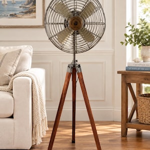 May include: A floor-standing fan featuring a circular metal cage with a wire grid design, four light-coloured blades, and a central circular emblem. The fan is mounted on a dark brown wooden tripod base with metal hardware and adjustable legs.