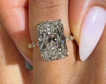 以下が含まれることがあります： ラディアントカットのダイヤモンドリングで、輝くファセット面を持つ大きな長方形のセンターストーンが特徴です。バンドは金属素材で作られており、シャンクに沿って小さな丸いアクセントストーンが並んでいます。OliverLabGrown。