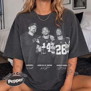 May include: A charcoal grey oversized t-shirt featuring a black and white graphic print of three men, identified as Campbell, Amon-Ra St. Brown, and Jahmyr Gibbs, with their names and signatures printed below each figure in a vintage style.