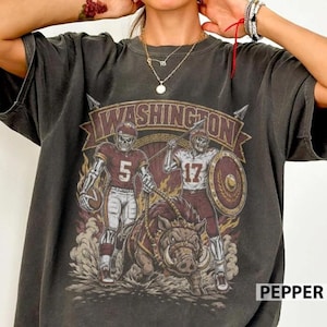 May include: A charcoal grey short-sleeve t-shirt featuring a graphic print of two skeleton football players and a wild boar. The text WASHINGTON is arched above the figures, with the numbers 5 and 17 displayed on the players' jerseys.