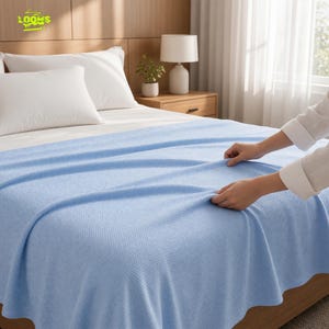May include: A light blue waffle-weave blanket spread across a bed. The fabric features a distinct grid-like texture throughout its surface. The blanket is draped over white pillows and a mattress, showcasing its soft, lightweight appearance.