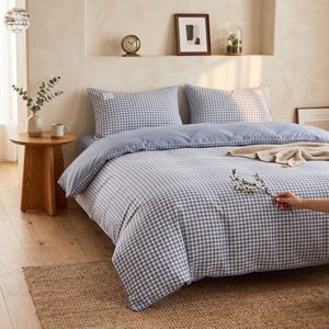 Pode incluir: Um conjunto de roupa de cama com padrão xadrez vichy azul e branco na capa de edredão e duas fronhas retangulares a condizer. O conjunto inclui uma dobra superior azul clara sólida e uma manta bege texturizada sobre a cama.