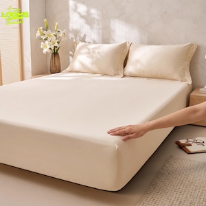 May include: A solid cream-colored fitted sheet covering a mattress, accompanied by two matching cream-colored pillowcases. A glass vase containing white lilies sits on a wooden nightstand next to the bed.