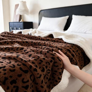 Puede incluir: Una manta de felpa con un estampado de leopardo marrón y negro en un lado y un forro de forro polar sherpa blanco sólido en el reverso. La tela suave y texturizada está colocada sobre una cama con almohadas blancas y un cabecero negro.