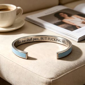 May include: A silver-toned metal cuff bracelet featuring a light blue enamel inlay on the exterior and the engraved black text "This too shall pass, BUT FUCKING HELL" along the inner surface of the band.
