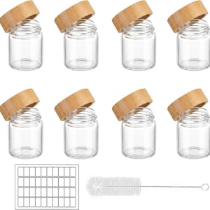 May include: A set of eight clear glass jars with cylindrical bodies and wide openings, each paired with a natural light brown wooden lid. The set includes a rectangular sheet of blank white labels and a long, thin wire-handled cleaning brush.