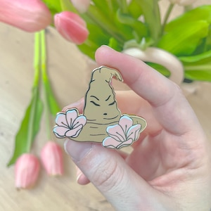 Peut inclure: Une broche en émail de couleur beige en forme de chapeau pointu et plié avec un visage aux yeux fermés et une bouche, ornée de deux fleurs rose clair avec des reflets blancs sur les côtés gauche et droit du bord, bordée de métal doré.