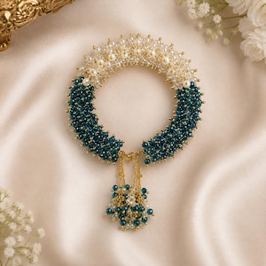 Puede incluir: Una pulsera circular de cuentas con una sección superior de perlas blancas y cristales transparentes, que pasa a una sección inferior de cuentas de color verde azulado profundo. Un colgante de cadena dorada con cuentas azules y blancas.