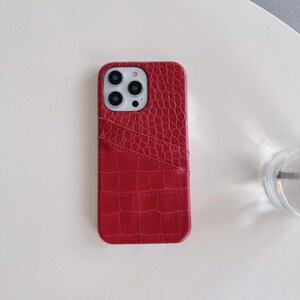 Puede incluir: Una funda para smartphone de color rojo con un patr&oacute;n texturizado de piel de cocodrilo. La funda cuenta con una ranura diagonal en la parte posterior para tarjetas y un recorte rectangular en la esquina superior izquierda para las c&aacute;maras.