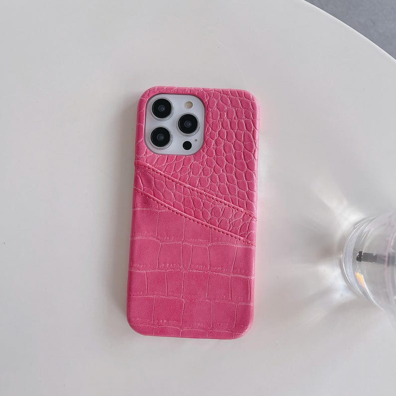Puede incluir: Una funda para smartphone de color rosa brillante con una textura de piel de cocodrilo. La funda incluye una ranura diagonal en la parte posterior para tarjetas y un recorte preciso para el sistema de c&aacute;mara trasera de tres lentes.