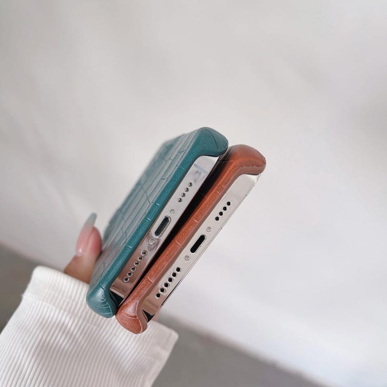 Puede incluir: Dos fundas para smartphone con un patr&oacute;n texturizado de piel de cocodrilo, una en color verde azulado profundo y la otra en un tono marr&oacute;n claro c&aacute;lido. Ambas fundas tienen recortes precisos para el puerto de carga y los altavoces.