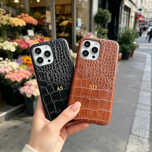 May include: Two smartphone cases with a textured crocodile skin pattern, one in black and one in brown. Each case features a card slot pocket and gold embossed initials, with AS on the black case and SD on the brown case, and a camera cutout.