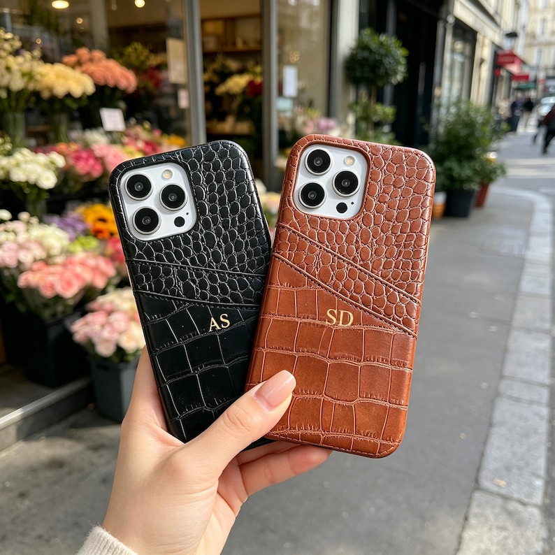 Puede incluir: Dos fundas para smartphone con un patr&oacute;n de piel de cocodrilo texturizado, una negra y otra marr&oacute;n. Cada funda cuenta con un bolsillo para tarjetas e iniciales doradas en relieve, AS en la negra y SD en la marr&oacute;n, y un recorte para la c&aacute;mara.