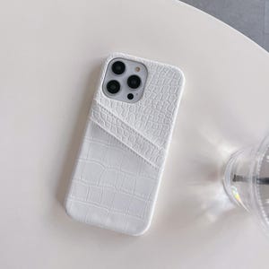 Puede incluir: Una funda para smartphone de color blanco con un patr&oacute;n texturizado de piel de cocodrilo. La funda incluye un bolsillo integrado para tarjetas en la parte posterior con una abertura diagonal. El &aacute;rea de la c&aacute;mara est&aacute; abierta y muestra tres lentes.