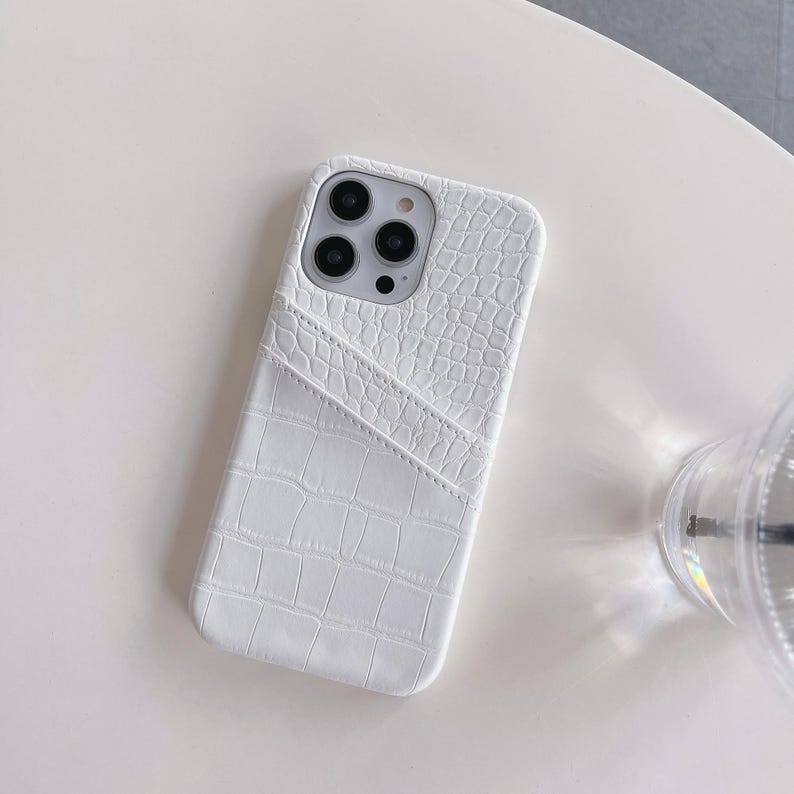 Puede incluir: Una funda para smartphone de color blanco con un patr&oacute;n texturizado de piel de cocodrilo. La funda incluye un bolsillo integrado para tarjetas en la parte posterior con una abertura diagonal. El &aacute;rea de la c&aacute;mara est&aacute; abierta y muestra tres lentes.