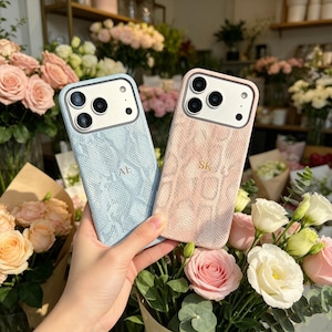 May include: Two smartphone cases with a textured snakeskin pattern, one in light blue with the initials AE and one in soft pink with the initials SK. Both cases feature a white camera cutout area with three circular lens openings and a flash.