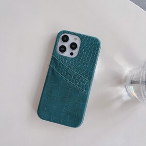 Puede incluir: Una funda de tel&eacute;fono color verde azulado con un patr&oacute;n texturizado de piel de cocodrilo y un bolsillo diagonal para tarjetas en la parte posterior. La funda presenta un recorte cuadrado para la c&aacute;mara con tres aberturas circulares y flash.