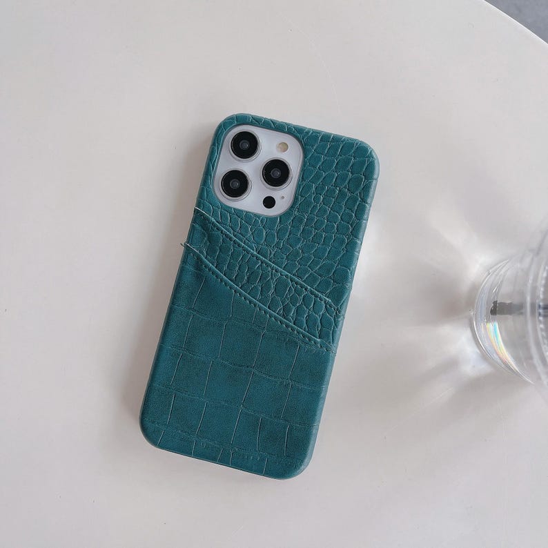 Puede incluir: Una funda de tel&eacute;fono color verde azulado con un patr&oacute;n texturizado de piel de cocodrilo y un bolsillo diagonal para tarjetas en la parte posterior. La funda presenta un recorte cuadrado para la c&aacute;mara con tres aberturas circulares y flash.