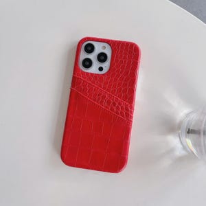 Puede incluir: Una funda para smartphone de color rojo brillante con un patr&oacute;n de textura de piel de cocodrilo. La funda incluye una ranura diagonal en la parte posterior para guardar tarjetas y un recorte preciso para la triple c&aacute;mara y el flash.